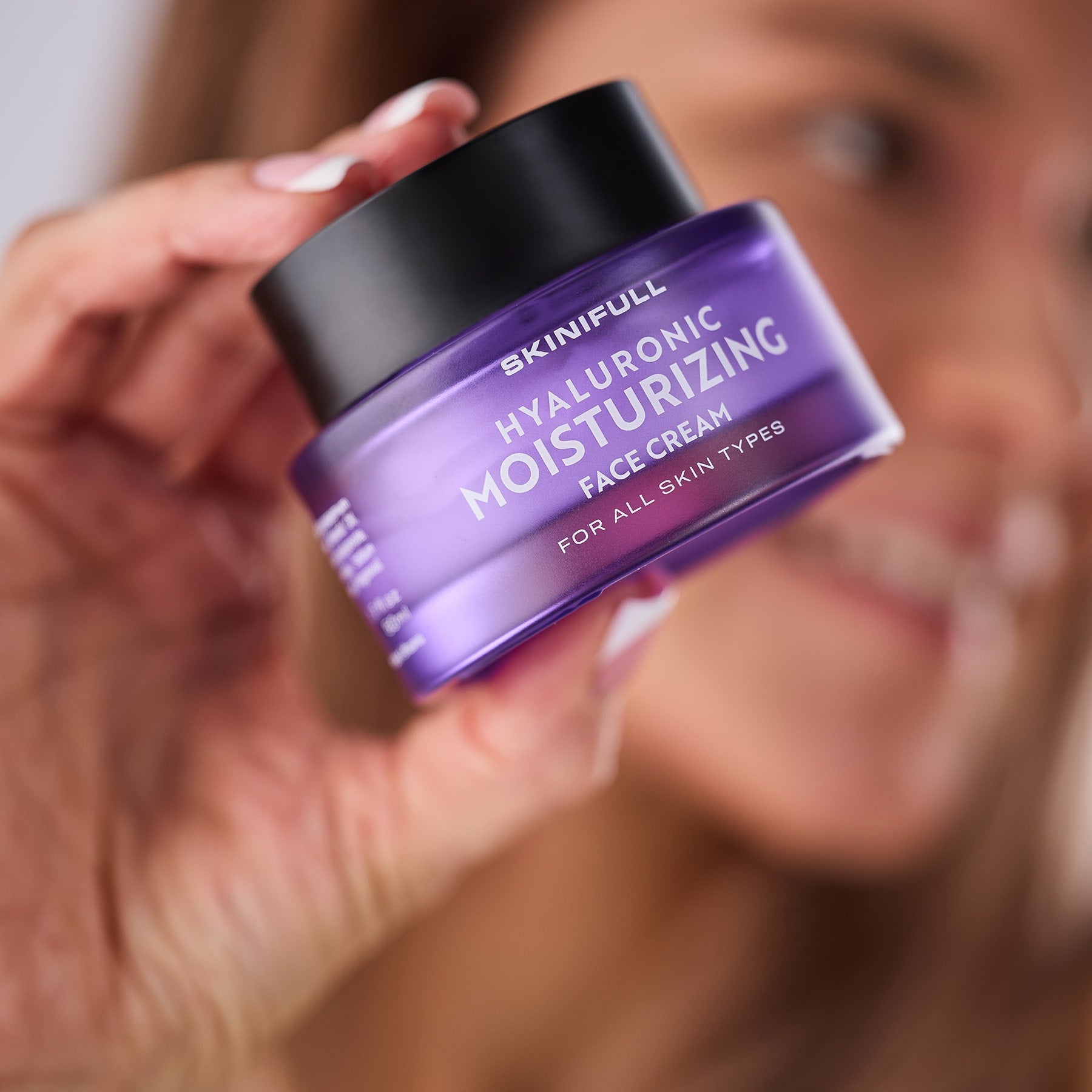 Moteris rankoje laiko Skinifull Hyaluronic Moisturizing Face Cream stiklainėlį, matosi šypsena fone