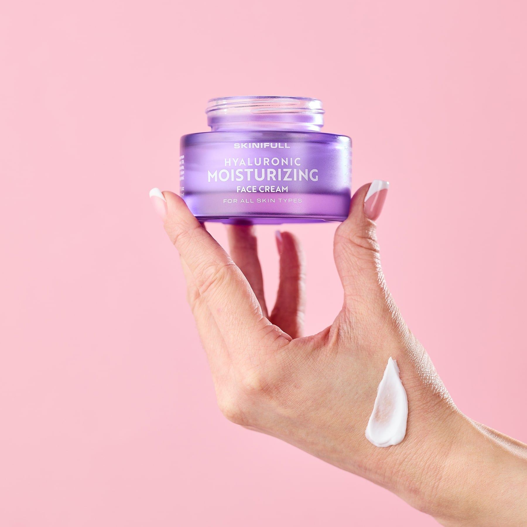 Skinifull Hyaluronic Moisturizing Face Cream stiklainėlis laikomas rankoje, ant odos užteptas kremo pavyzdys, rožiniame fone